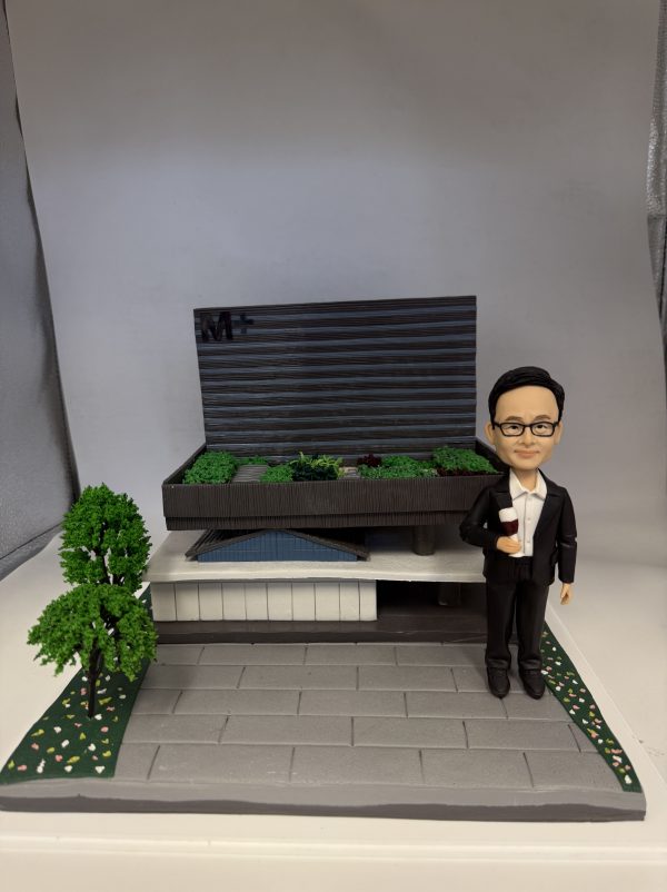 (客製化「西裝男士與M+博物館建築」紀念公仔 Q版男士公仔（身穿黑色西裝套裝、白色襯衫，配戴黑框眼鏡的成熟男士，右手持一杯紅酒，左手自然垂下，表情溫和自信，展現專業與優雅的氣質） 微縮建築場景（背景為精緻的現代建築模型，頂部有顯眼的「M+」標誌，呈現深色橫條紋外牆與綠化屋頂設計，建築下方有白色結構與藍色細節；底座為灰色石磚鋪面，左側有一棵立體綠樹，兩側邊緣點綴綠草與小白花） 專業形象、建築落成、慶祝時刻、現代藝術、成熟穩重 商務禮品、建築師紀念、落成典禮贈禮、退休紀念、辦公室擺飾)