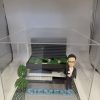 一座企業主題 diorama：右側是身著黑色西裝與白襯衫、戴眼鏡的人像公仔，右手握小獎盃；中央至後景為層次分明的灰黑色建築模型，屋頂佈置綠植區塊與步道；前方地面以灰磚鋪設，放置醒目藍色“SIEMENS”立體字，左前方有樹與花草點綴，整體封裝於透明壓克力盒中。