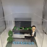 一座企業主題 diorama：右側是身著黑色西裝與白襯衫、戴眼鏡的人像公仔，右手握小獎盃；中央至後景為層次分明的灰黑色建築模型，屋頂佈置綠植區塊與步道；前方地面以灰磚鋪設，放置醒目藍色“SIEMENS”立體字，左前方有樹與花草點綴，整體封裝於透明壓克力盒中。