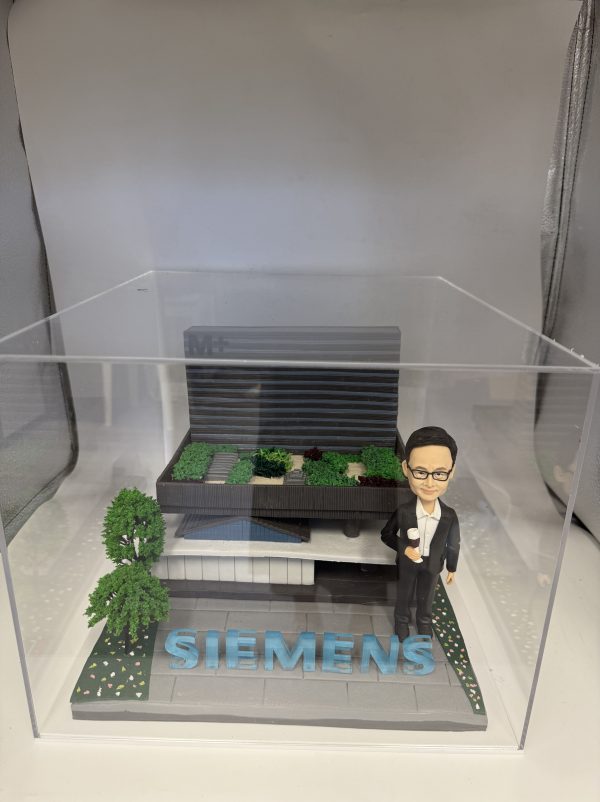 一座企業主題 diorama：右側是身著黑色西裝與白襯衫、戴眼鏡的人像公仔，右手握小獎盃；中央至後景為層次分明的灰黑色建築模型，屋頂佈置綠植區塊與步道；前方地面以灰磚鋪設，放置醒目藍色“SIEMENS”立體字，左前方有樹與花草點綴，整體封裝於透明壓克力盒中。