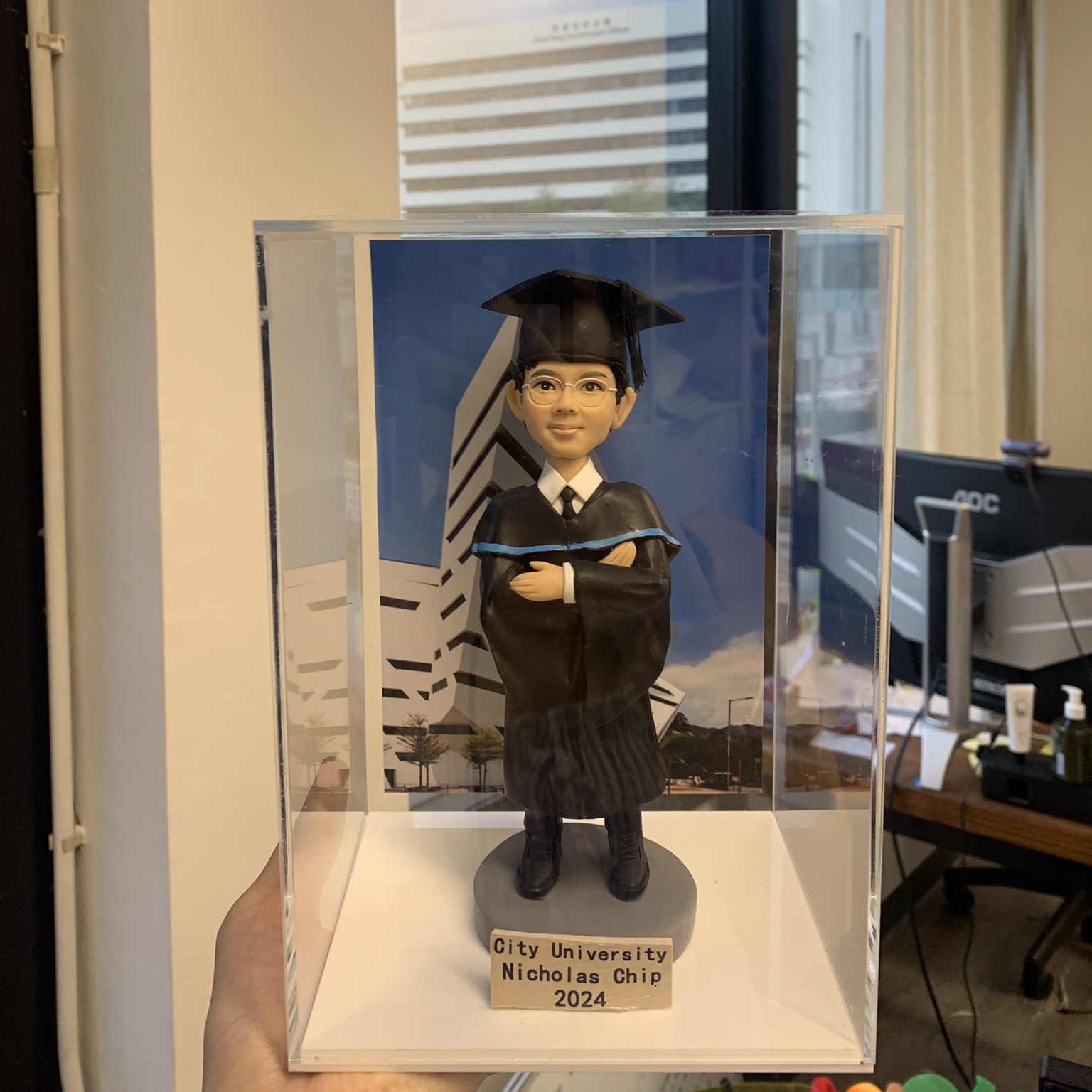 (客製化「香港城市大學」畢業男生公仔與展示盒 Q版畢業男生公仔（身穿黑色學士袍，袍身有細緻的褶皺紋理，領口處有藍色滾邊設計，內搭白色襯衫與黑色領帶。頭戴黑色學士帽與流蘇，佩戴銀框眼鏡，面帶自信微笑，雙手交叉抱胸，展現出沉穩與專業的氣質） 精緻展示盒與背景（公仔被安置在一個透明亞克力展示盒中，防塵且美觀。背景是一張印有現代化白色幾何建築的照片，推測為香港城市大學的標誌性建築——邵逸夫創意媒體中心，增強了畢業紀念的場景感） 專屬紀念銘牌（公仔站立於灰色圓形底座上，前方設有長方形木色銘牌，印有黑色文字：「City University / Nicholas Chip / 2024」，清楚標示了學校名稱、畢業生姓名及畢業年份） Q版寫實風、畢業禮物、香港城市大學、CityU、建築背景、亞克力展示盒、客製化公仔、眼鏡男生 大學畢業紀念、校友禮品、辦公桌擺件、送給兒子的畢業禮物、學術成就慶祝)