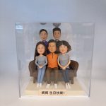 一組家庭主題的人像公仔擺設放在透明展示盒內：前排三人坐在咖啡色沙發上，左側長髮女生與右側短髮女生穿灰色上衣與運動鞋，中間男子穿橙色上衣與眼鏡；後排兩位站在後方，一位穿紫色上衣、一位穿深色上衣。底座前方有淺色小牌寫著「媽媽 生日快樂！」。背景是簡潔的室內白色環境，展示盒邊緣與反光可見。