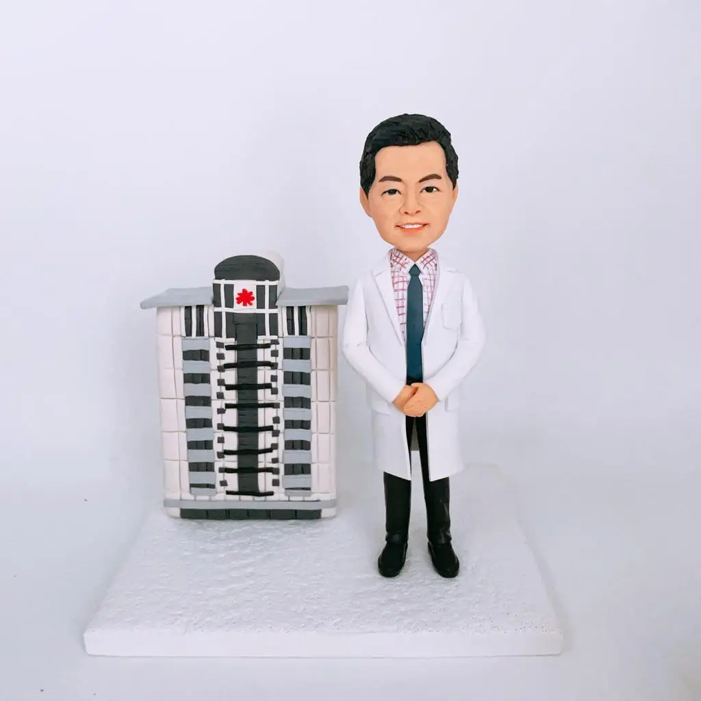 白色底座上的手作模型：左側是一棟縮尺的明愛醫院外牆立體建築，採米白與深灰配色，中央有深色垂直玻璃帶與層層陽台線條，頂部圓拱位置嵌有紅色十字花形標誌；右側站著一位醫師造型的人像公仔，短髮微笑，穿白袍、格紋襯衫與藍色領帶，雙手於身前交握，黑長褲與皮鞋。背景為純白布景，強調醫院建築元素與醫護主題。