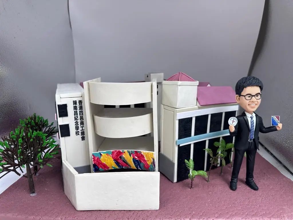 分割式「立體建築對比圖」。左側（BEFORE）：香港四邑商工總會陳南昌紀念中學校舍外觀，立面以紅、藍、黃等豔色立柱節奏排列，入口處有英文校名牌與學生聚集的街景。右側（AFTER）：手作 diorama 模型，粉紅色地面上重現校園一隅：左側為白色弧形走廊與開放式平台，外緣以多彩筆觸裝飾；中段是白牆結合方形塔樓與粉紅屋頂；右側為黑色窗格教學樓並加上淺藍水平線條，周圍點綴小型綠植。畫面右邊站著一位商務造型的人像公仔，短髮、戴眼鏡、穿黑色西裝打領帶，手持圓形徽章與小卡，展現學校主題與紀念氛圍。