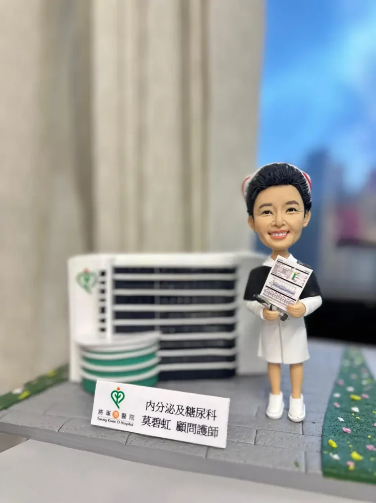 分割式「立體建築對比圖」。左側（BEFORE）：晴天藍天下的將軍澳醫院外觀，白色與綠色窗帶的層疊建築量體，立面有圓角轉折與豎向窗格，主樓上可見心形綠色院徽。前景是行道樹與道路圍欄。右側（AFTER）：手作 diorama 擺設，灰色磚紋底座上重現醫院外牆的白色弧形樓翼與深色水平窗帶，左側圓柱體量與綠白相間環形層次；前方放置白色立牌印有「將軍澳醫院」與部門與姓名字樣。右側站立一位護理主題的人像公仔，髮髻與紅白髮箍，穿黑白相間的制服與白色便鞋，手持文件夾與表單，微笑面容，旁邊鋪有綠色花植步道，背景為室內淺景深。