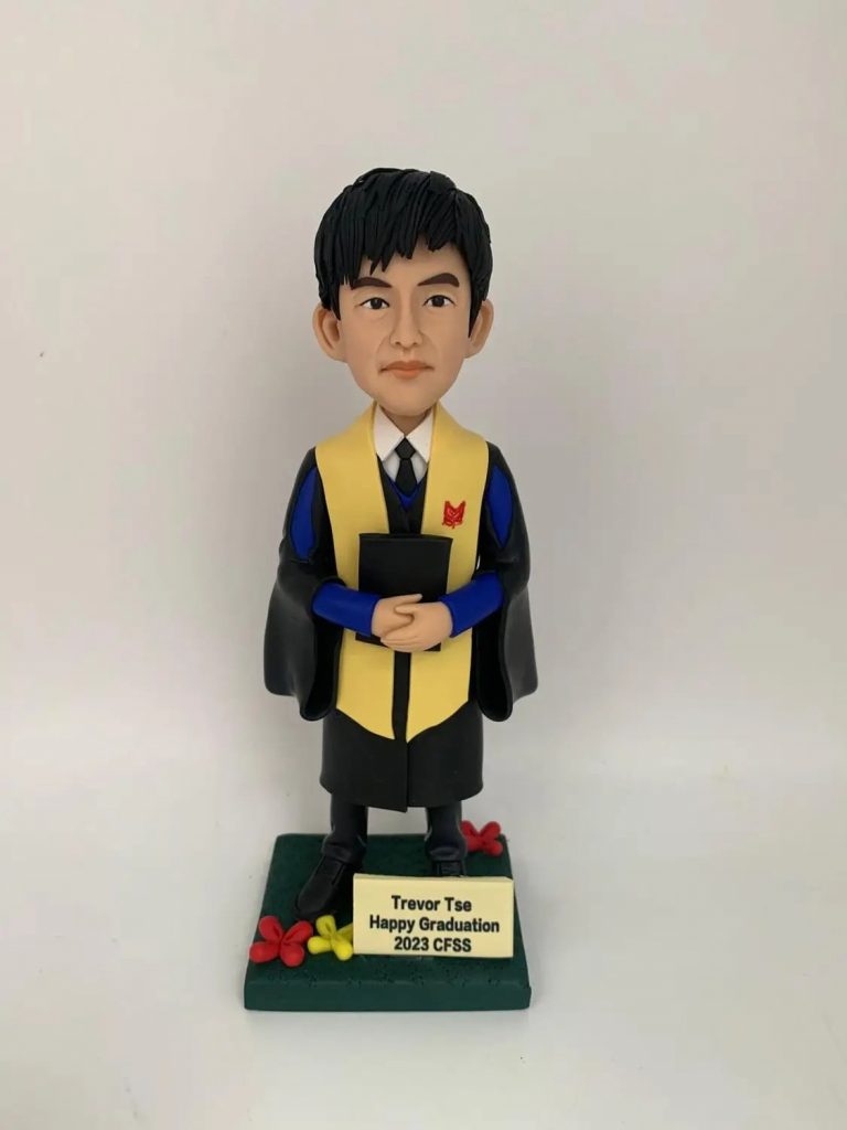 (客製化 CFSS 畢業紀念公仔) 這是一份慶祝 Trevor Tse 於 2023 年從 CFSS 畢業的專屬紀念禮物。 服飾細節：公仔身穿黑色學士袍，搭配顯眼的黃色領巾 (Stole) 及藍色長袖內襯，左胸處印有紅色校徽，內搭白襯衫與黑領帶，色彩層次分明。 動作神態：雙手交疊手持黑色證書夾（或文件夾）於胸前，表情自信沉穩，展現畢業生的端莊氣質。 底座設計：綠色方形底座象徵草地，點綴著紅黃色的小花裝飾。前方名牌清晰印有「Trevor Tse」、「Happy Graduation」及「2023 CFSS」字樣，充滿祝福寓意。