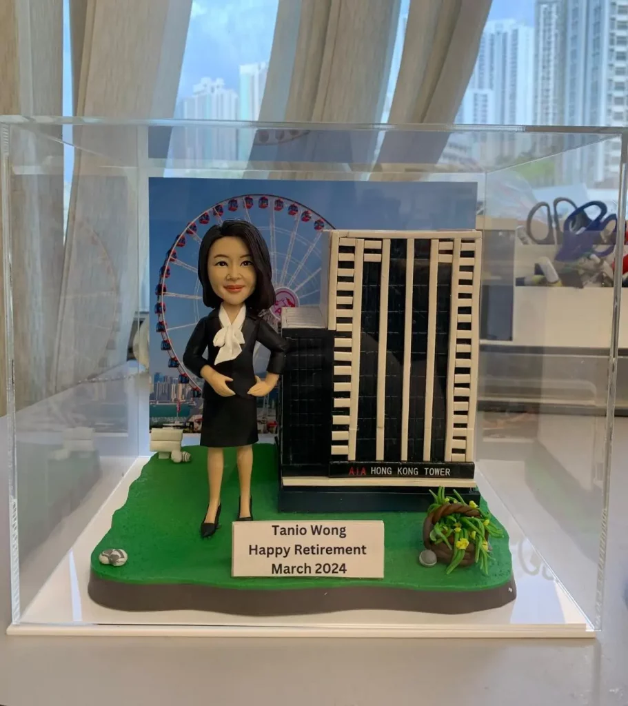 透明展示盒內的客製場景：一個穿黑色套裝、白色領結襯衫的女性人像公仔站在綠色草地底座上，雙手叉腰微笑。她旁邊是兩棟縮尺大樓模型，其中一棟立面有黑白直條並標示「HONG KONG TOWER」，前方草地角落有小石燈與植物裝飾。背景板印有摩天輪與城市天際線。底座前方銘牌寫著「Tanio Wong Happy Retirement March 2024」。整體置於亞克力盒內，周圍可見室內窗簾與辦公文具的景深。
