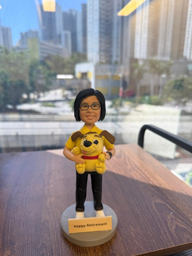 一款單人主題的人像公仔站在灰色圓形底座上，前方附小牌寫有「Happy Retirement」。角色留中短黑髮、戴黑框眼鏡，穿黃色上衣、黑色長褲與白鞋，手錶配件在左手腕。雙臂抱著一隻黃色狗狗造型的布偶玩具（紅色項圈、棕色耳朵）。作品擺放在木質桌面上，背景是窗外的高樓城市景與樹木，日光明亮。