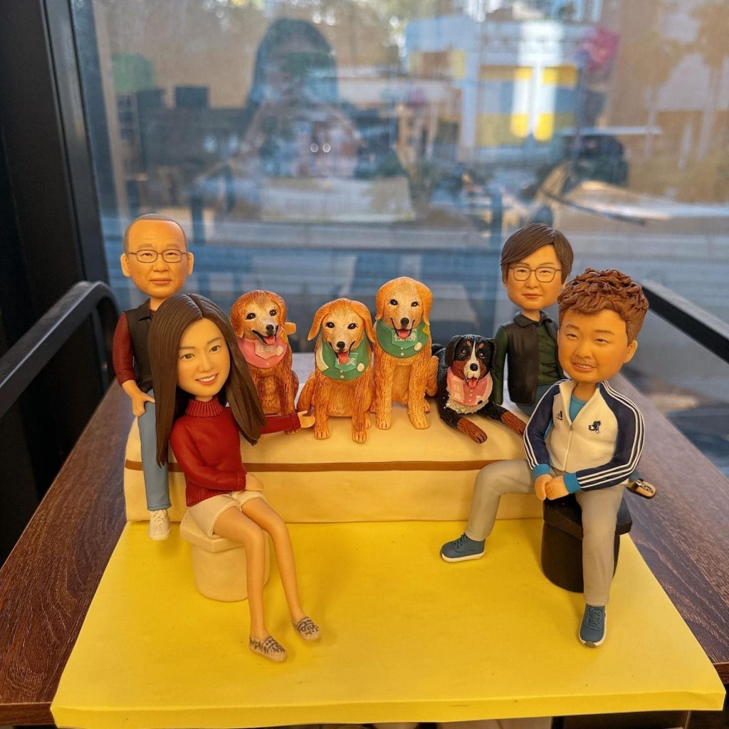 一組全家福人像公仔與狗狗公仔的迷你場景:四位角色圍坐在階梯式座台兩側與前方,中央台階上並排四隻狗狗,包括三隻金色長毛犬配綠色與粉色領巾,以及一隻黑白棕配色的犬隻戴粉紅掛牌。人物服裝各異:左側為紅色上衣與白色短裙、後方站著穿背心的長者;右側為藍白運動外套與運動鞋的年輕人,旁邊還有一位戴眼鏡的角色。整體置於黃色底板與木質桌面上,背後是窗景。