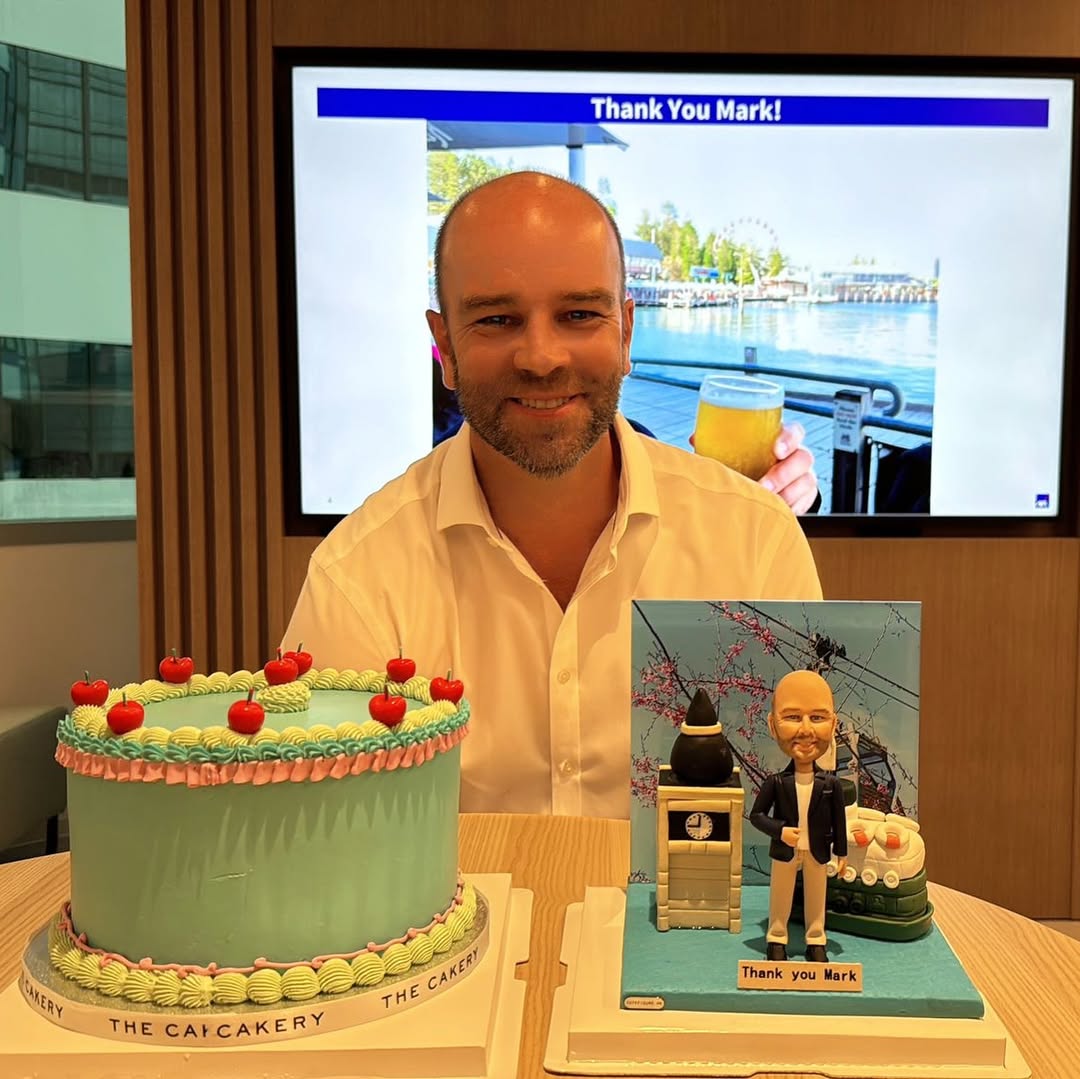 藍色地台上站立一位穿深藍外套、淺色長褲與黑鞋的人像公仔，前方小牌寫著「Thank you Mark」。旁邊有小鐘樓造型、黑白圓錐物與綠白色船體元素，背景板印有櫻花枝與纜車電線的照片。場景置於淺木桌上。