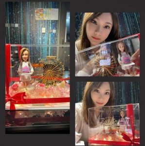三張合成照片展示同一份禮物：透明展示盒內放置一款女性人像公仔，身穿淺色洋裝、手捧粉紅生日蛋糕；背景有金色摩天輪裝飾，底部鋪滿粉色花材與暖色燈串。展示盒外以紅色緞帶與標籤綁飾，上寫「Forever friend」。旁邊可見拍照者與禮盒的合照，場景在室內、背板為深色與亮點紋理。