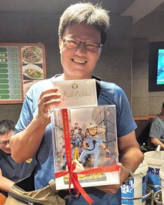 室內合照中，一位男子微笑手持透明展示盒與祝福卡片。展示盒以紅色緞帶綁繩，內有工程主題的人像公仔：身穿淺藍工作服與白色安全帽，腰間掛對講機，站在黃黑相間的地面警示區旁，前方有黃色圍欄與路錐。背景板印有多位穿藍色制服的人員與貨櫃場景，盒底前方附有小銘牌。