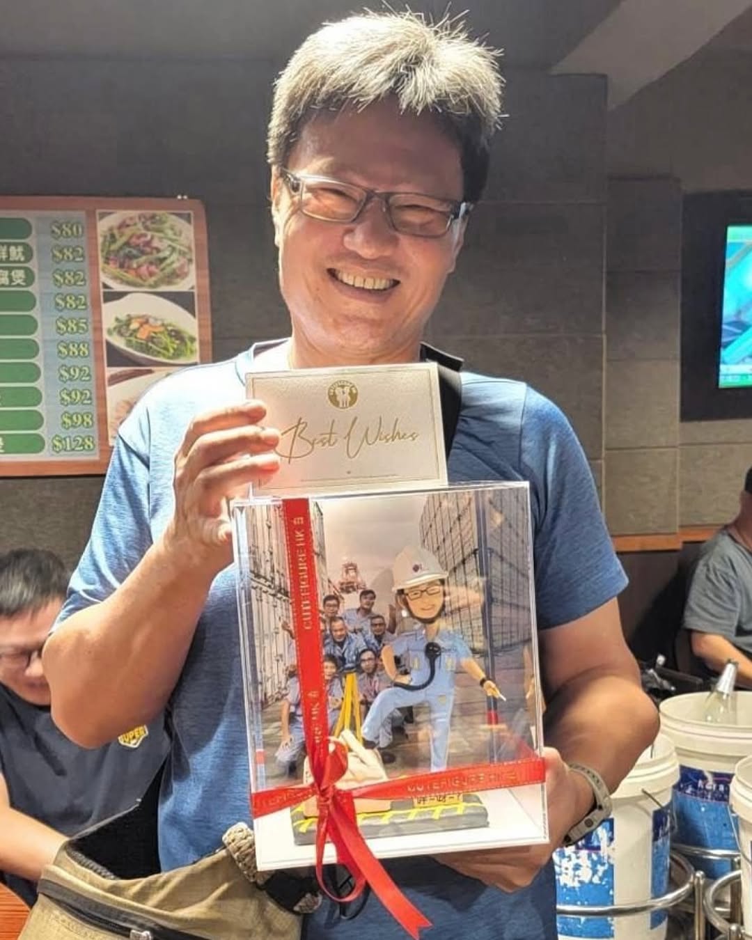 室內合照中，一位男子微笑手持透明展示盒與祝福卡片。展示盒以紅色緞帶綁繩，內有工程主題的人像公仔：身穿淺藍工作服與白色安全帽，腰間掛對講機，站在黃黑相間的地面警示區旁，前方有黃色圍欄與路錐。背景板印有多位穿藍色制服的人員與貨櫃場景，盒底前方附有小銘牌。