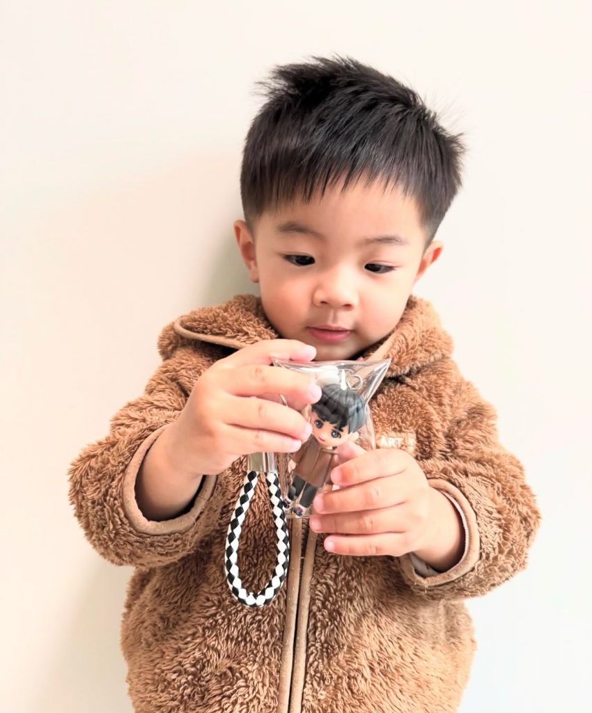 近距離視角，小朋友雙手高舉透明吊袋，袋中可見同款Q版人像公仔與金屬圈扣，毛絨外套質感清晰。