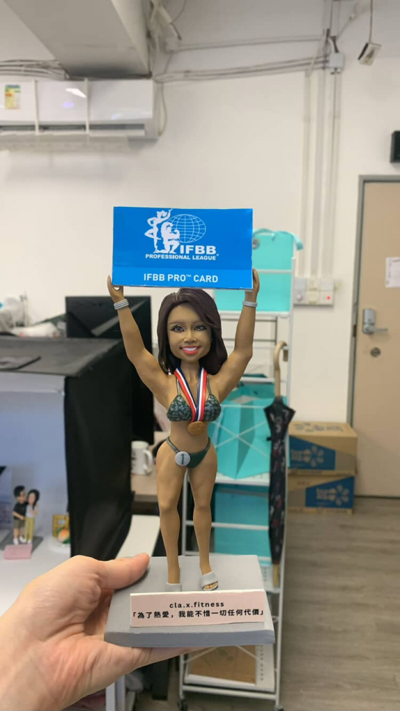 (客製化「IFBB Pro 健美選手」榮耀時刻公仔 Q版健美選手公仔（女性角色身穿墨綠色比基尼賽服，展現健美緊實的肌肉線條與自信體態，膚色呈現健康的古銅色，雙手高舉藍色的「IFBB PRO CARD」職業卡，脖子上掛著紅藍白配色的獎牌，長髮披肩，臉上洋溢著獲勝的喜悅與驕傲笑容） 勵志語錄底座（灰色方形底座，穩重簡約，前方立牌印有IG帳號「cla.x.fitness」以及勵志金句「為了熱愛，我能不惜一切任何代價」，深刻傳達選手對夢想的堅持與付出） Q版寫實風、肌肉線條刻畫、職人手作、榮耀時刻、夢想成真 健美比賽紀念、職業卡獲獎禮物、健身教練贈禮、勵志收藏、個人成就紀錄)