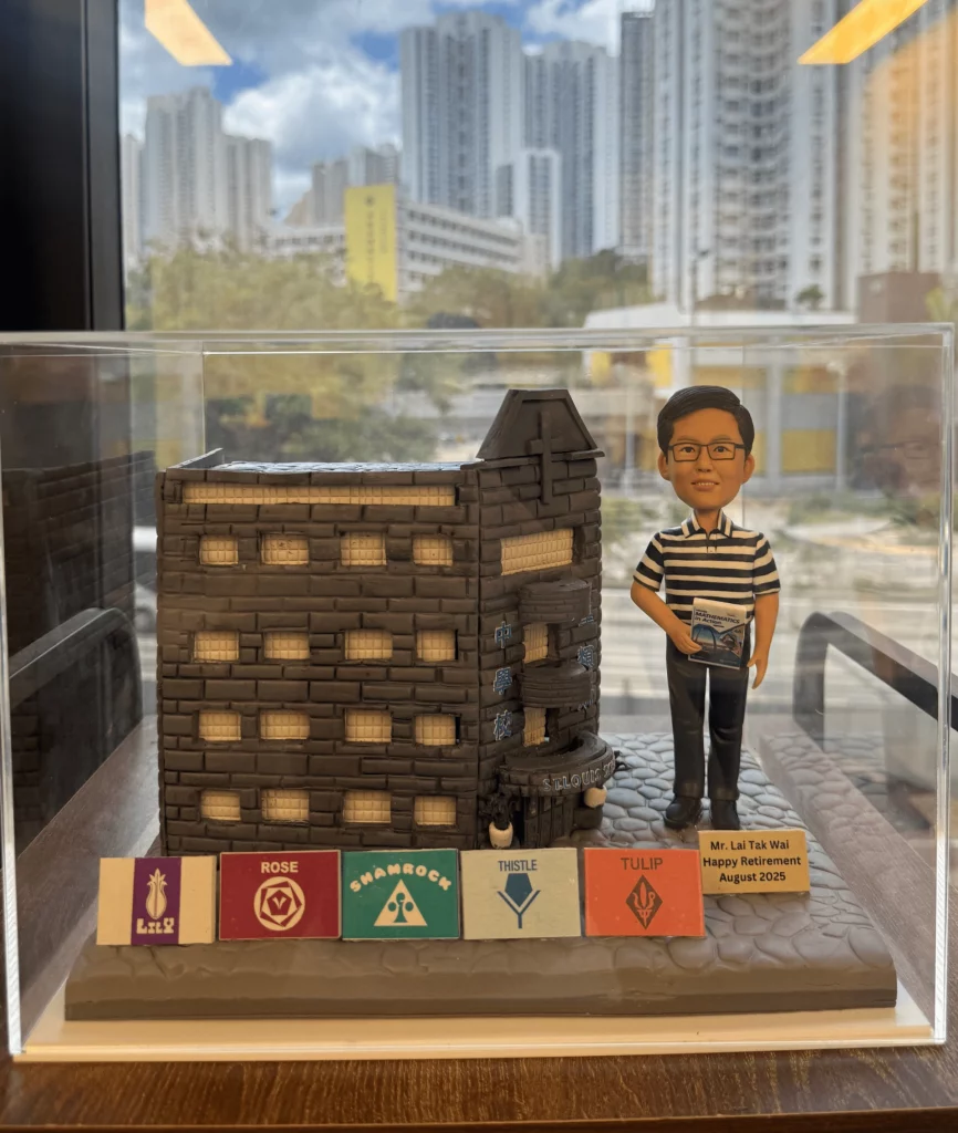 分割式「立體建築對比圖」。左側（BEFORE）：聖類斯中學的街角老校舍仰視照片，灰褐色外牆、轉角圓弧陽台與連續方窗，入口位於轉角處，周圍是密集高樓。右側（AFTER）：透明展示盒內的手作 diorama，還原深灰磚塊質感的校舍量體與轉角三層圓弧平台，牆上以藍字寫有「聖類斯中學」，入口門楣標示 ST. LOUIS SCHOOL；前景設有代表不同學社的彩色徽章牌。右側站立一位戴眼鏡、穿黑白橫間 Polo 衫與長褲的人像公仔，手持數學教材，旁邊附有「Happy Retirement」小牌，整體呈現溫馨致敬的校園氛圍。