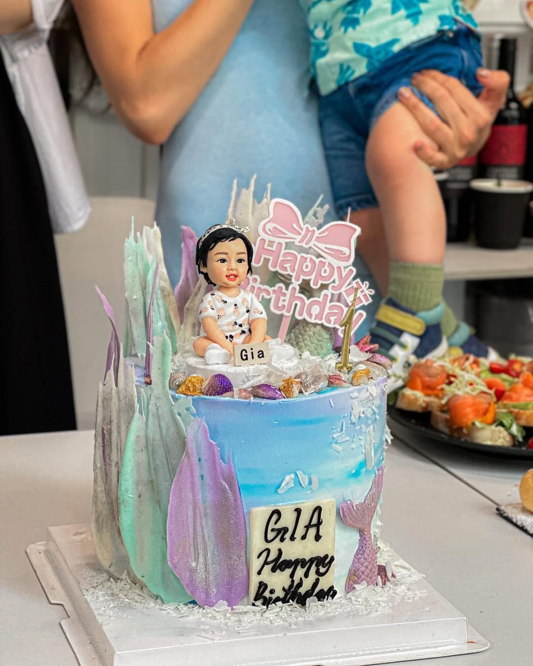 藍紫漸層海洋風生日蛋糕，上方坐著嬰幼兒造型的人像公仔，戴小皇冠、穿白底花紋衣，前有「Gia」小姓名牌與金色數字1蠟燭；蛋糕四周以貝殼、海星、紫色魚尾和晶片狀裝飾點綴，側面寫有「GIA Happy Birthday」。背景可見大人雙手與餐具。