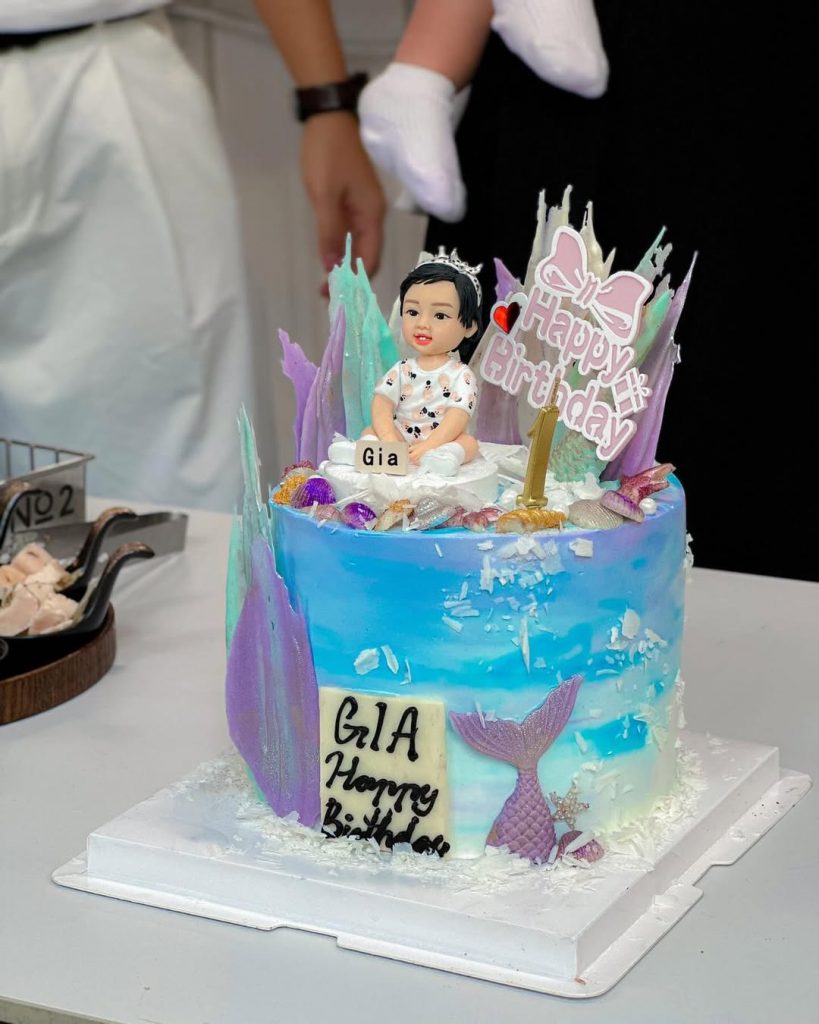 客製化Q版女寶寶公仔蛋糕——夢幻美人魚海洋主題 手工人物公仔與蛋糕細節 （圖片展示了一個精緻的生日蛋糕，頂端坐著一個Q版女寶寶黏土公仔。寶寶留著黑色短髮，頭戴銀色小皇冠，身穿印有粉色圖案的白色連身衣，雙手捧著寫有名字「Gia」的小方牌，表情天真可愛，眼神靈動。蛋糕體以藍色和白色奶油抹面，營造出漸層的海洋波浪質感。裝飾細節豐富，包括紫色與藍綠色的直立糖片（彷彿珊瑚或海浪）、散落的貝殼造型裝飾，以及蛋糕側面立體的紫色美人魚尾巴。頂部插著粉紅色的「Happy Birthday」蝴蝶結插牌以及金色的數字「1」蠟燭，側面還有一塊手寫「GIA Happy Birthday」的白巧克力牌，整體設計充滿童話色彩。） 環境背景與光影 （蛋糕放置在白色的桌面上，背景處於室內環境，畫面後方模糊可見站立的大人（其中一人似乎抱著小孩），左側有裝著餐具的籃子及些許食物，顯示這是在一個熱鬧的生日派對或宴會現場。現場光線明亮均勻，讓蛋糕上的藍紫色調顯得夢幻且吸睛，公仔作為視覺焦點清晰突出。） 軟陶公仔、蛋糕裝飾、週歲蛋糕、美人魚主題、海洋風、女寶寶、皇冠造型、客製化公仔、Gia、生日派對 成長紀念、甜蜜祝福、童話色彩、獨家訂製、派對焦點、溫馨時刻、充滿愛意、夢幻氛圍