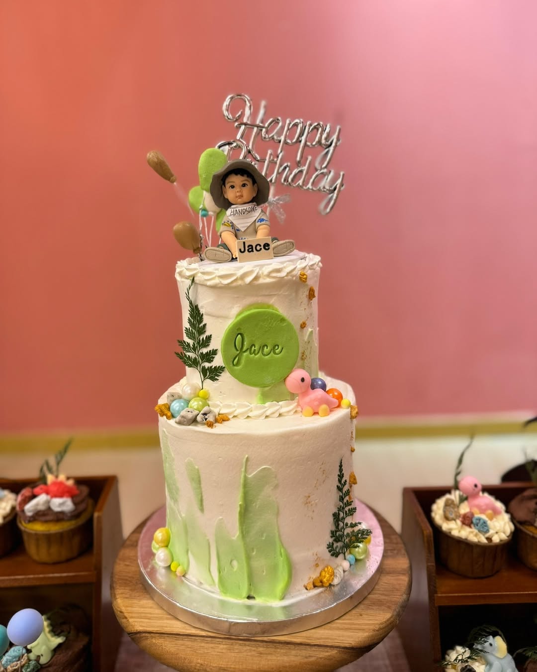 兩張照片展示同一款生日蛋糕：一顆綠白配色的雙層奶油蛋糕，側面以抹刀綠色筆觸、葉材與珍珠糖球點綴；上層蛋糕頂端坐著一個嬰幼兒造型的人像公仔，戴寬邊帽、穿淺色上衣與綠色短褲，腳穿運動鞋，手前方有「Jace」名牌。後方插有銀色「Happy Birthday」字樣與綠、棕色氣球裝飾。蛋糕下方木托盤與旁側甜點台可見小鴨、彩色球等裝飾。