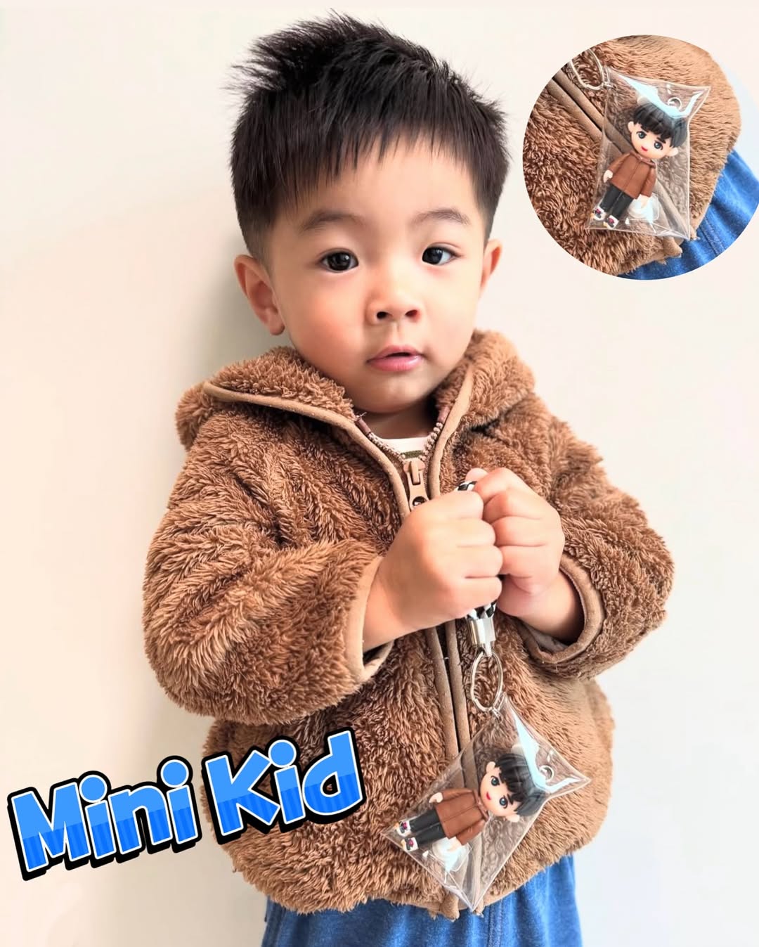 一位小朋友穿著棕色毛絨拉鍊外套與藍色下裝，雙手握著透明軟殼吊袋內的Q版人像公仔鑰匙圈；公仔黑色短髮、穿棕色外套與深色褲、白鞋，搭配黑白編織手繩與金屬鑰匙圈。左下角以藍色粗體字寫「Mini Kid」，右上角有放大圓框特寫，展示同款公仔的細節。
