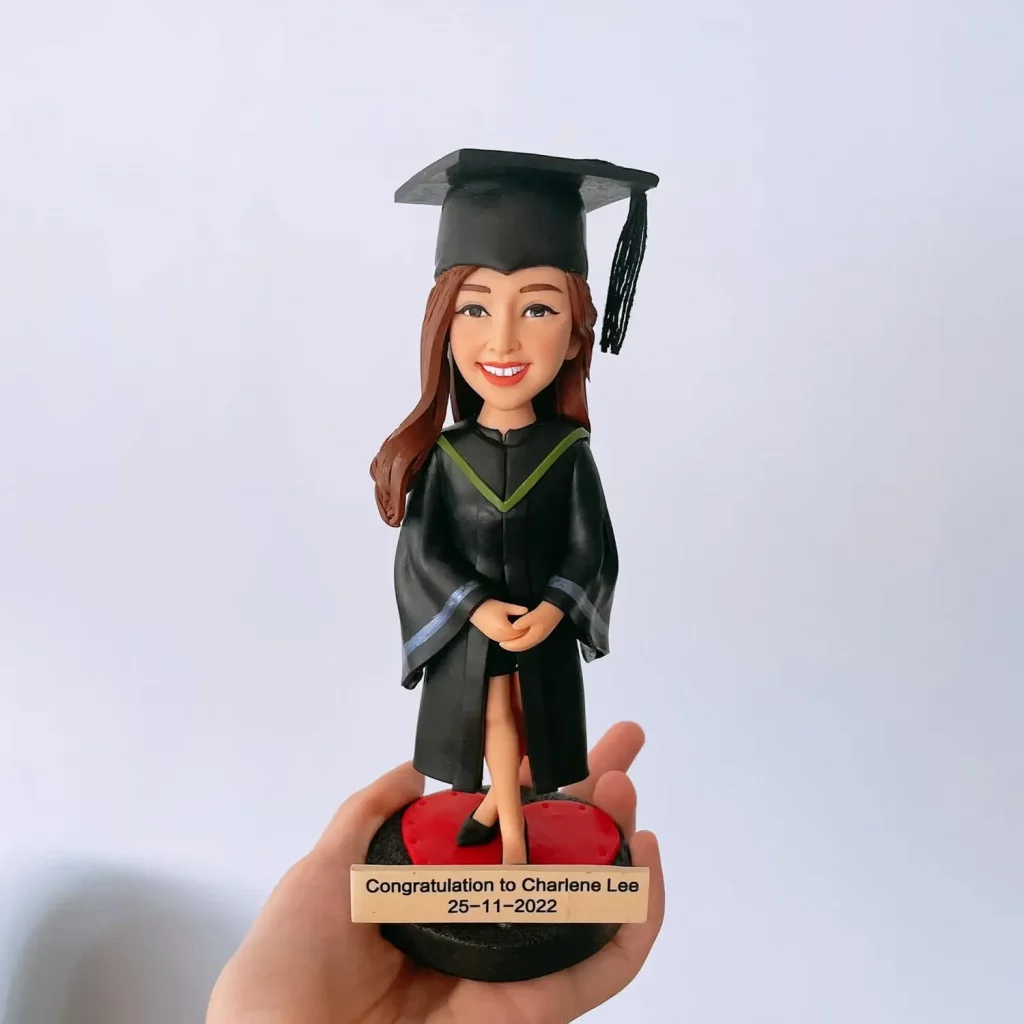一款以香港中文大學風格為靈感的人像公仔：戴黑色學士方帽與黑色流蘇，穿黑色畢業袍，胸前為深灰V形披肩配綠色細邊，袖口帶藍灰飾條。公仔雙手於身前交握、站在紅黑圓形底座上，底座名牌刻有祝賀字樣與日期。背景為簡潔的淺色牆面，手掌托住底座展示。