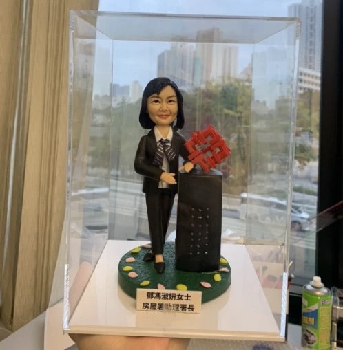 透明壓克力展示盒中的桌面擺設：一位穿黑色西裝與條紋領帶的人像公仔，微笑站在綠色圓形草地底座上，邊緣點綴彩色卵石；右側是一座黑色長方柱，頂部插有紅色幾何齒輪狀立體標誌。前方白色銘牌以中文標示職銜，背景可見窗外城市景與高樓。