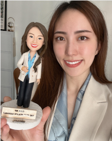 一位女性手持與自身穿搭相似的人像公仔,背景在室內。公仔為長髮造型,穿米色外套、藍色上衣與深色長褲,站在白色圓形底座上;底座前方有小金屬牌,刻有「ELKA NIP」與「THE BEST BRANCH MANAGER」等英文字樣。持公仔者穿米色外套與藍灰色絲巾,對鏡頭微笑。