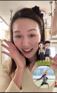 一位女性在室內對著鏡頭微笑並舉手示意，另一手拿著一座雙人人像公仔。公仔立於圓形綠色草地台座上：左側為短髮女公仔，穿無袖運動上衣與九分運動褲，赤腳站立；右側為短髮男公仔，穿無袖運動背心與短褲，亦赤腳站立。背景可見吊掛繩與瑜伽空中布等運動器材、天花板燈與窗簾。畫面右下角嵌有一個圓形小圖，顯示一對男女在戶外進行雙人瑜伽（Acro Yoga）動作，城市天際線作為背景。