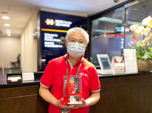 室內銀行櫃檯前的合照：一位穿紅色短袖制服與識別證、配戴口罩與眼鏡的人士，雙手捧著一個綁有紅色緞帶的透明展示盒。盒內是一尊銀行職員主題的人像公仔，公仔也穿紅色上衣並站立於方形台座上。背景可見深色木質櫃檯、電子顯示牌、宣傳立牌與花卉擺設，呈現銀行分行環境。