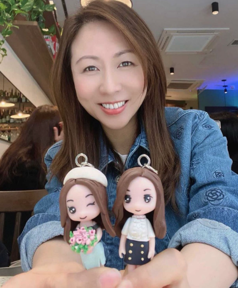 一位坐在咖啡廳環境中的女性對著鏡頭微笑，穿牛仔外套，雙手在鏡頭前方托著兩個Q版人像公仔鑰匙圈。左側公仔戴白色貝雷帽、單眼眨眼微笑，手捧粉綠色系花束，穿淡綠洋裝；右側公仔披長髮，穿白色上衣與海軍藍釦飾短裙，表情可愛呆萌。背景可見吊掛植栽、餐廳燈飾與其他座位，呈現「客人回圖」的實拍分享氛圍。