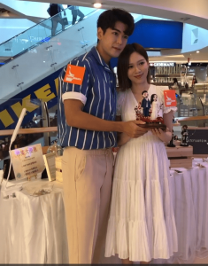 商場活動現場，兩位KOL在展桌前合照，手持一組客製人像公仔作品。公仔為成對角色：一位穿深色西裝、另一位著白色禮服，置於帶紅褐色枝葉裝飾的底座上。背景可見手扶梯、品牌招牌，以及桌上宣傳牌、收納盒與其他公仔展示物，屬推廣或路演情境。