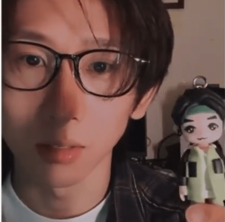 兩張畫面組合的截圖：近景中，一位佩戴黑框眼鏡的人士面向鏡頭，畫面右側靠近鏡頭處舉著一個Q版人像公仔鑰匙圈；公仔有黑髮、綠色頭帶與淺綠拼接外套，頭頂連接金屬吊環。另一畫面顯示同一人士坐在沙發前，以單手拿起同款迷你公仔端詳，畫面下方有中文字幕，桌面擺放小型植栽與日常物件，呈現分享或開箱情境。
