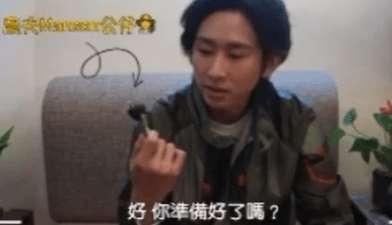 兩張畫面組合的截圖：近景中，一位佩戴黑框眼鏡的人士面向鏡頭，畫面右側靠近鏡頭處舉著一個Q版人像公仔鑰匙圈；公仔有黑髮、綠色頭帶與淺綠拼接外套，頭頂連接金屬吊環。另一畫面顯示同一人士坐在沙發前，以單手拿起同款迷你公仔端詳，畫面下方有中文字幕，桌面擺放小型植栽與日常物件，呈現分享或開箱情境。