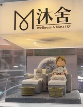 多張照片展示同一套開張人像公仔：一位穿著淺灰藍浴袍的Q版人物坐在按摩足浴沙發上，腳前有絨面腳墊與兩個木質足浴桶，旁邊另置一張同款空沙發與托盤小茶杯。背景板為灰色圓形圖案，整體置於灰白色地台。作品被放入透明燈箱中，燈箱上印有「沐舍 Wellness & Massage」字樣，固定在店舖外牆作為開張或門面擺設；遠景可見店鋪招牌、圓形窗與街景。
