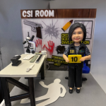 一座以 法證鑑識（CSI） 為主題的個人化人像公仔作品。 主角身穿黑色 CSI 制服背心，胸前印有識別徽章，手持「證物10號」標示牌，神情自信而親切。 身後背景板模擬「CSI ROOM」刑事現場調查室，牆上印有手槍、彈殼、血手印與偵查標誌等元素。 場景桌上擺放顯微鏡、鍵盤、指紋工具與法證記錄冊，地上則有白色人形輪廓貼示，完整重現真實辦案氣氛。 整體配色以灰、黑、紅為主，象徵冷靜與專業，亦突顯法證工作者的細心與責任感。 作品為送給團隊法證成員的特製 職場榮譽禮物， 以立體造型致敬在第一線默默付出的 CSI 專業精神。