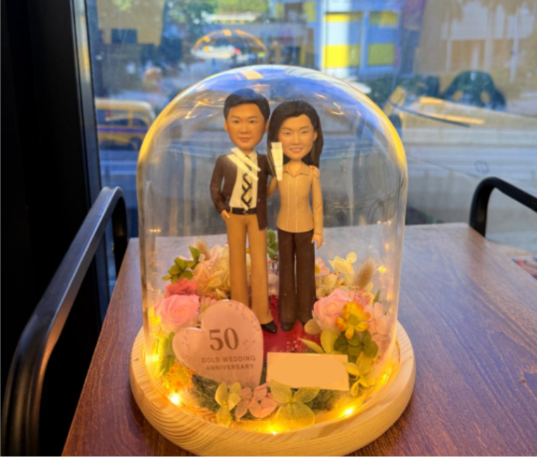 一款為慶祝 結婚50週年(金婚紀念) 而特製的雙人 人像公仔擺設。 作品以溫馨真摯的夫妻形象為主角,人物姿態自然、表情柔和,展現半世紀相守的默契與溫度。 底座鋪滿粉紅與象牙色系的仿真花材,象徵愛情長久與幸福綻放。 前方擺放粉色心形石牌,上刻「50 GOLD WEDDING ANNIVERSARY」,紀錄珍貴裡程碑時刻。 整體覆以透明玻璃罩,內圈點綴柔黃燈帶,營造溫暖浪漫的氛圍。 此設計是贈予父母、長輩或伴侶的 金婚紀念人像禮物, 以手工雕塑的形式永恆記錄「愛與時間」的故事。