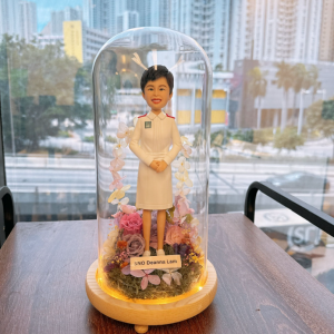 這款作品以 護士（Nurse）專業形象 為靈感，展現溫暖與堅毅的護理精神。 人物身穿整齊的白色護士制服，肩襯紅邊徽章細節清晰；短髮造型搭配明亮笑容，傳達出專業、親切與關懷。 底座採用乾燥花園設計，融合粉、紫、米白色花材，象徵溫柔守護與生命綻放。 作品外層覆以透明玻璃罩，並以暖黃燈圈環繞，營造出柔亮而典雅的紀念氛圍。 前方名牌可客製姓名或銜頭，使每個細節都更具專屬感。 此公仔作品非常適合作為 退休紀念、晉升賀禮、或感謝醫護人員辛勞的專業禮物， 以手工藝術方式致敬「白衣天使」無私奉獻的精神與微笑背後的力量。