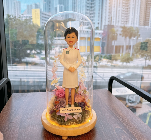 這款作品以 護士（Nurse）專業形象 為靈感，展現溫暖與堅毅的護理精神。 人物身穿整齊的白色護士制服，肩襯紅邊徽章細節清晰；短髮造型搭配明亮笑容，傳達出專業、親切與關懷。 底座採用乾燥花園設計，融合粉、紫、米白色花材，象徵溫柔守護與生命綻放。 作品外層覆以透明玻璃罩，並以暖黃燈圈環繞，營造出柔亮而典雅的紀念氛圍。 前方名牌可客製姓名或銜頭，使每個細節都更具專屬感。 此公仔作品非常適合作為 退休紀念、晉升賀禮、或感謝醫護人員辛勞的專業禮物， 以手工藝術方式致敬「白衣天使」無私奉獻的精神與微笑背後的力量。