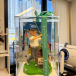 這款作品以熱愛高爾夫的紳士為主題，完美呈現 揮桿瞬間的專注與從容氣度。 人物身穿專業高爾夫球裝，配搭印有品牌標誌的球帽與球桿， 神情自信爽朗，動作流暢自然，將揮桿動態定格於最佳角度。 腳下綠地草坪與球洞細節刻畫精緻，旁邊配置個人姓名立牌與球具包， 整體色調清新明亮，背景結合球場視覺設計，營造出活力十足的休閒氛圍。 外層透明展示盒搭配品牌綠色緞帶包裝，既具收藏價值亦適合送禮展示。 此作品特別適合作為 退休日禮、生日賀禮、或高爾夫球友專屬紀念公仔， 以立體藝術方式紀錄那份投入、沉穩與熱愛運動的生活品味。