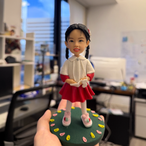 這款作品以 小女孩的天真笑容與甜美氣質 為主題，展現童年的純粹與快樂。 女孩綁著整齊的雙辮，髮梢點綴小花飾，臉上帶著自然明亮的微笑； 身穿紅白相間的洋裝與粉色鞋子，色彩搭配柔美可愛，充滿活潑朝氣。 雙手輕握玩具小兔，腳下站立在點綴花瓣的綠色圓形底座上， 象徵童年花園般的夢幻世界。作品線條細膩流暢， 不論正視或側看，都能感受到童趣與溫馨氛圍。 這款人像公仔特別適合作為 生日禮物、兒童節紀念、親子留念或成長里程收藏品， 讓最可愛的笑靨以立體藝術形式永遠保存，成為心中最溫暖的時光印記。