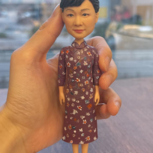 這款作品以 旗袍女性 為主題，展現東方女性的溫婉與高雅氣質。 人物神情柔和自信，髮髻盤起整齊端莊，微笑中流露出溫暖親切的風度。 她身穿一襲 深紫紅色碎花旗袍，細緻的花卉紋理與布料質感皆以手工上色刻畫， 完美重現傳統服飾的婉約之美與時代韻味。 腳踏紫色高跟鞋，立姿挺拔自然，整體氣場沉靜又不失優雅。 作品配有簡約白色底座，突顯人物主體之美，適合長久擺設與收藏。 此公仔特別適合作為 母親節禮物、長輩紀念公仔、榮退致敬禮或中式主題裝飾品， 以細緻雕工與藝術情感，傳遞敬意與感謝， 讓優雅姿態與溫柔笑容，永恆留存於歲月中。