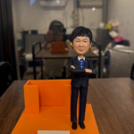 這款作品以 專業教師形象結合卡片座設計，展現知性氣質與實用美感。 人物身穿深色西裝，內搭藍色襯衫，雙手交叉、神情穩重， 體現教育者的自信與從容，傳遞“以德育人、以心啟智”的精神。 底座採用亮橙色設計，象徵活力與啟發， 附加的插槽結構可作為 名片、筆記、Memo 卡或小信件收納座， 在辦公桌或教職桌上既雅緻又具紀念意義。 前方金屬銘牌刻有「天道酬勤育桃李，倫才樹德啟眾心」， 為整體增添感恩與敬意的氣氛。 此設計特別適合作為 教師節禮物、榮退致敬、導師紀念或校慶贈禮， 將尊敬與謝意以藝術形式呈現， 讓教育之光持續溫暖人心