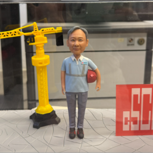 這款作品以 建築工程師 為主題設計，致敬多年專業堅守與匠心精神。 人物身穿工作制服，神情穩重親切，手持安全帽象徵責任與專業； 身後搭配迷你版 黃色吊塔模型 與 企業標誌牌， 完整重現建設現場的象徵性元素，極具職業代表意義。 底座以灰色地磚紋理設計，呼應工程基石的穩固與踏實， 整體封裝於透明展示盒中，既防塵又適合長久收藏。 前方金屬銘牌刻有「林利輝 榮休之樂」， 傳遞對敬業精神的尊敬與祝福。 此公仔特別適合作為 退休紀念、公務榮退、工程師表揚 或 建築業禮贈訂製， 以立體模型方式記錄職場成就， 讓每一道建築線條，化為人生精彩的印記。