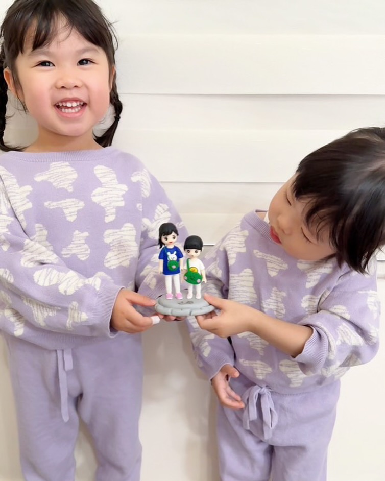 兩位穿淺紫套裝的小朋友並肩站在白色牆前，微笑握著一座雙人Q版人像公仔；公仔立於灰色圓形台座，上身分別為寶藍與白色T恤，胸前有兔子與黃色小鴨圖案，腳穿粉白運動鞋，兩人都斜背圓形綠色毛絨感側包。