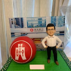 綠色磚紋台座上的桌面擺設：左側是一顆紅色球體，上有白色中文標記的圓形標誌；右側站著一位人像公仔，白襯衫、黑色長褲與棕色皮鞋，雙手叉腰微笑；後方背景板呈現恒生銀行門面與玻璃建築外觀，前側留有淺色銘牌位置。