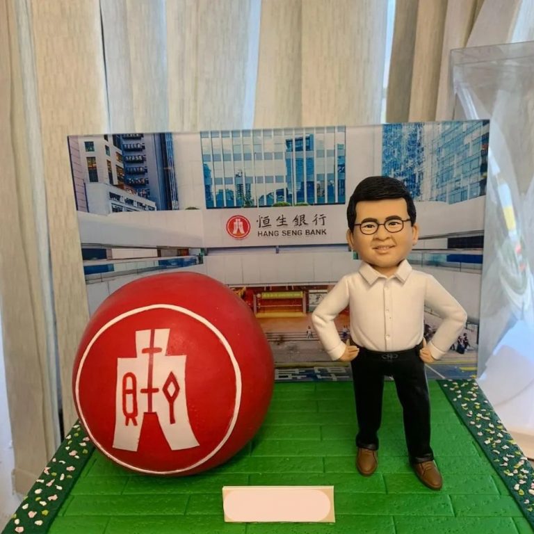 綠色磚紋台座上的桌面擺設：左側是一顆紅色球體，上有白色中文標記的圓形標誌；右側站著一位人像公仔，白襯衫、黑色長褲與棕色皮鞋，雙手叉腰微笑；後方背景板呈現恒生銀行門面與玻璃建築外觀，前側留有淺色銘牌位置。