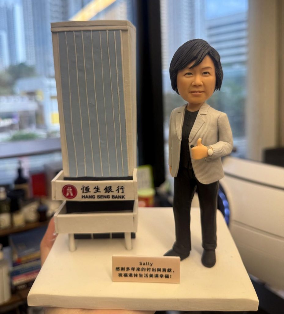 桌面擺拍的客製模型：白色底座上，左側是一棟簡化的高樓立體建築，立面為淺藍色並以多條豎向白線表現玻璃帷幕；樓前設有黑白相間的「恆生銀行 HANG SENG BANK」小型門頭牌，左側帶紅色圓形徽記。右側站著一位商務造型的人像公仔，短髮、穿灰色條紋西裝外套與黑色上衣長褲，腳穿深色皮鞋，右手比讚，左腕配戴手錶。底座前緣有金色小銘牌，印有“Sally”與感謝祝福字樣；背景為模糊的室外高樓窗景，整體呈現恆生銀行元素與個人化禮贈主題。
