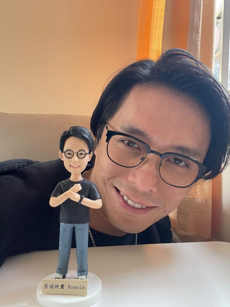 一位男士笑容滿面地與他訂造的Q版真人公仔合照。公仔穿黑色T恤、藍色褲和波鞋，戴圓框眼鏡和手錶，底座刻有「香城映畫 Ronnie」，是令人開心的訂造人像公仔。