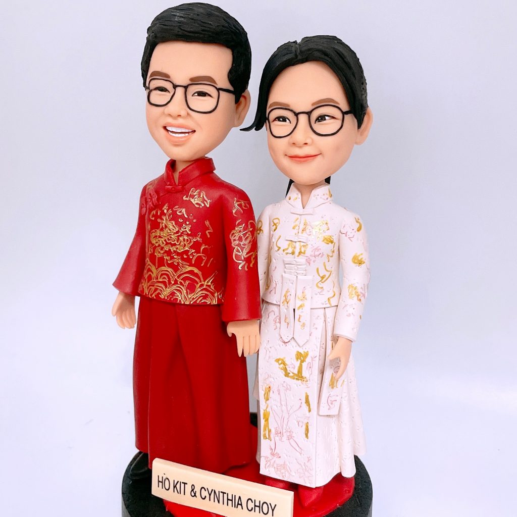 成對Q版公仔身穿中式禮服站立：男方著大紅唐裝繡金龍紋，女方穿白底金紋長衫裙，兩人皆配黑框眼鏡，站在黑色圓座上，前方名牌刻有姓名。