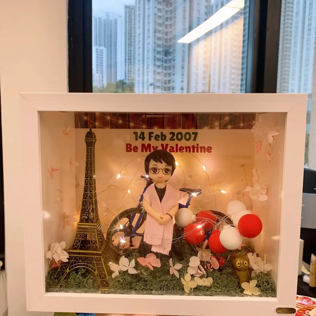 白色相框中的情人節主題場景：Q版人物站在藍色單車旁，周圍有紅白氣球與花材裝飾，左側為迷你艾菲爾鐵塔模型；背景寫有「14 Feb 2007 Be My Valentine」，並以暖白燈串點綴。